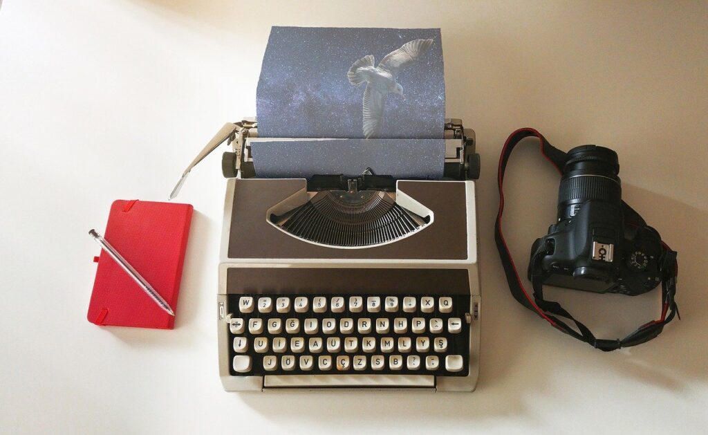 Picture of a journal and pen, typewriter with a picture of a bird, and a camera next to each other.