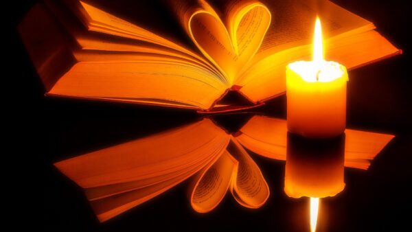 Photo of an open book with pages folded in the shape of a heart next to a lit candle.